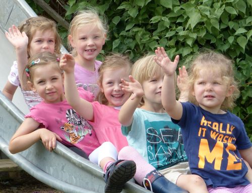 Sechs fröhliche Kinder sitzen auf einer Rutsche und winken in die Kamera.