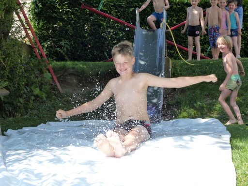 Junge rutscht auf einer Rutsche ins Wasser, Kinder spielen im Hintergrund.