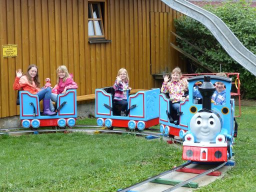 Fünf Kinder sitzen in bunten Zugwagen, einer fährt als Lokomotive.