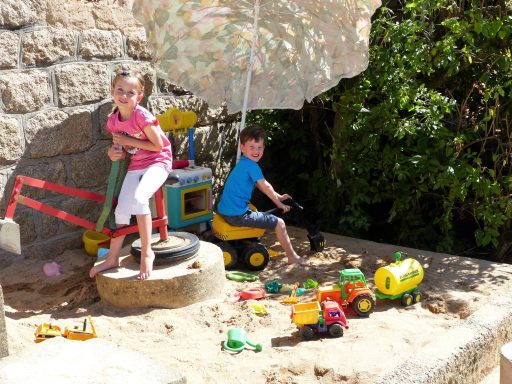 Zwei Kinder spielen mit Spielzeug im Sandkasten unter einem Sonnenschirm.