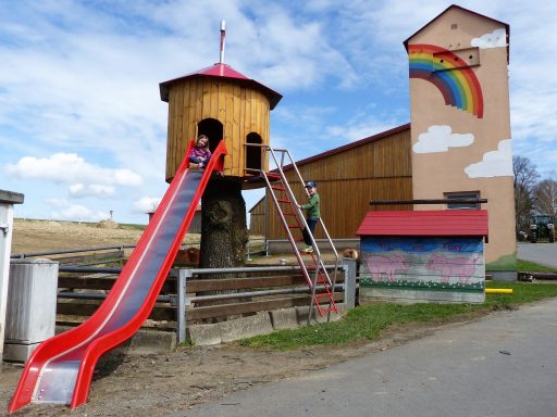 Spielplatz mit Baumhaus, Rutsche und buntem Wandbild von einem Regenbogen.