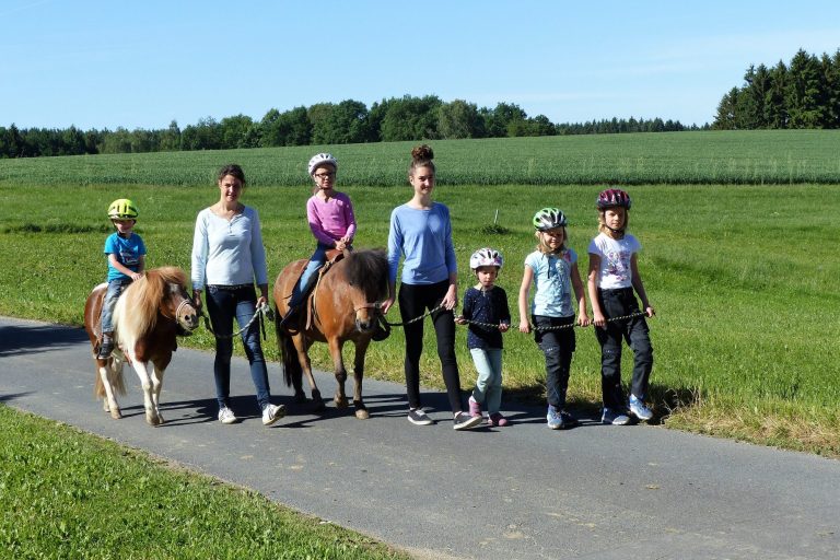 Gruppe von Kindern und Erwachsenen mit Ponys auf einem Weg in einer grünen Landschaft.